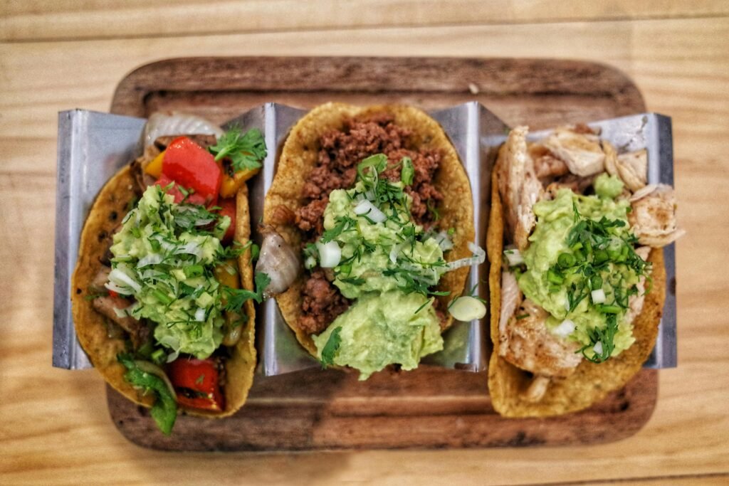 Three vibrant tacos with fresh guacamole, veggies, and meat on a wooden tray.